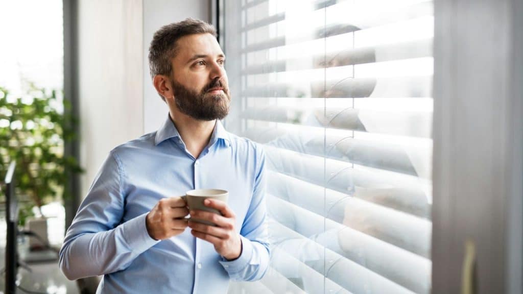 A bearded man in a blue shirt is standing by a window with blinds, holding a mug.