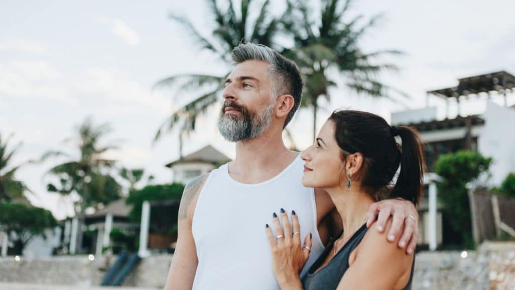 A silver-haired, bearded man and a woman are standing outside with palm trees in the background.