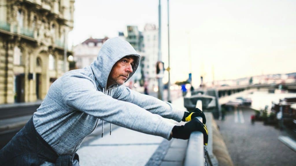 A serious man in a gray hoodie and gloves is leaning on a railing in a city.