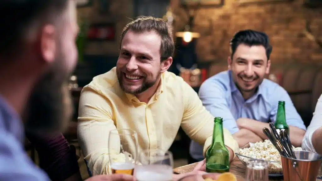 A happy bearded man in a yellow shirt is drinking beer with a group of friends.