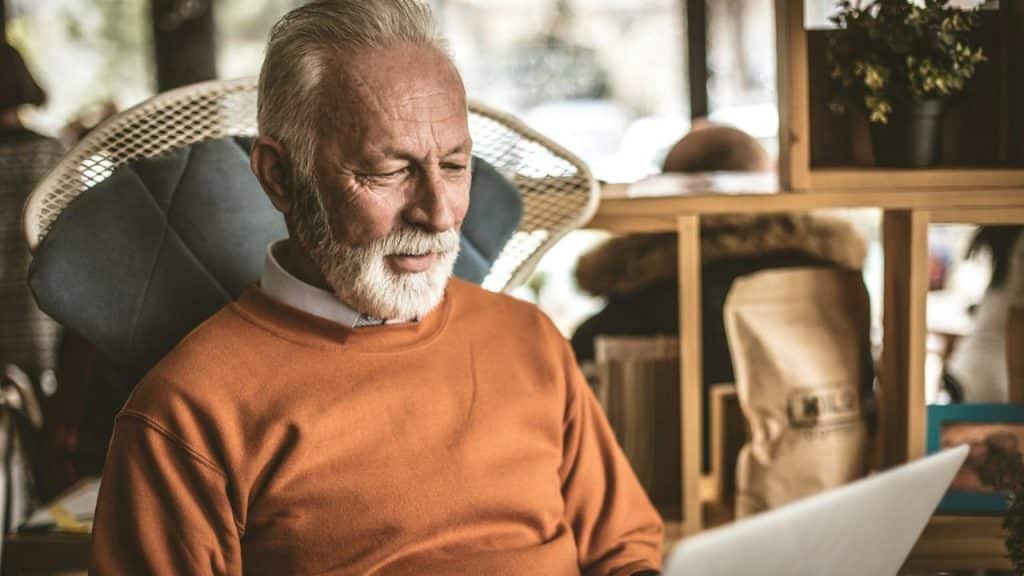 An older, silver-haired man with a white beard is looking at a laptop while sitting in a chair.