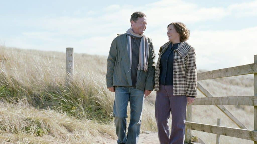 A couple is smiling at each other while walking on a grassy, windy path.