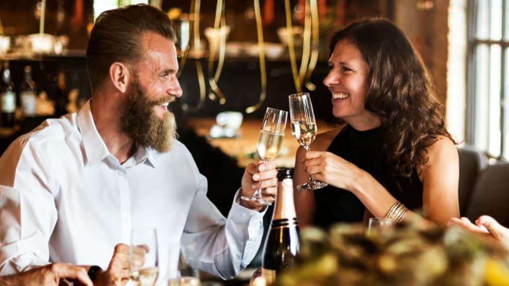 Bearded man and woman smiling at each other while clinking champagne glasses on a date.
