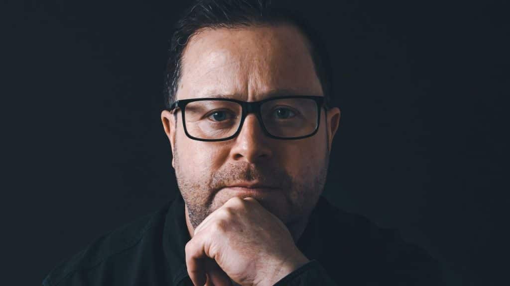Close-up portrait of a man with glasses and a beard, resting his chin on his hand.