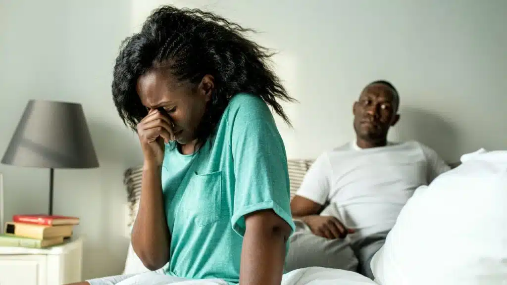 A distressed woman in a teal shirt sits on a bed, holding her hand to her face, while a man sits blurred in the background.