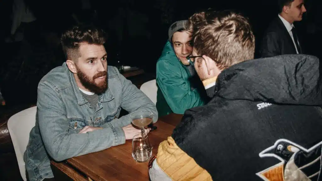 Three men sit around a dark wooden table in a dimly lit setting, possibly a bar, talking.