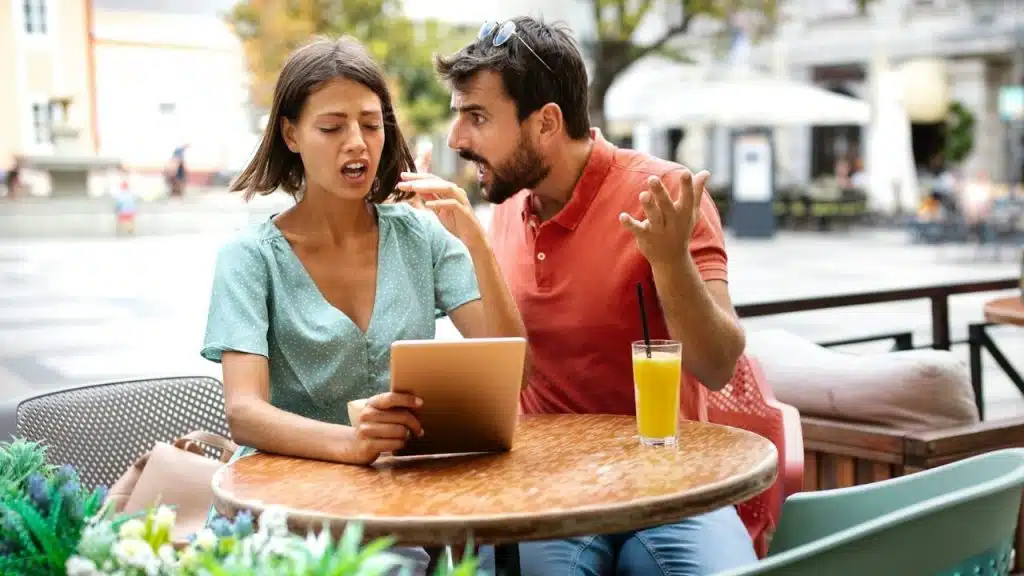 A woman holds a tablet while a distressed man in an orange polo shirt argues with her at an outdoor cafe.