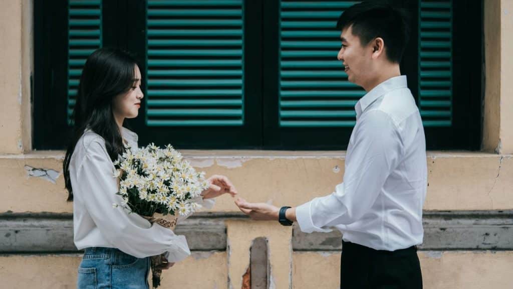 A man and woman in white shirts stand facing each other; the woman holds a bouquet of daisies.