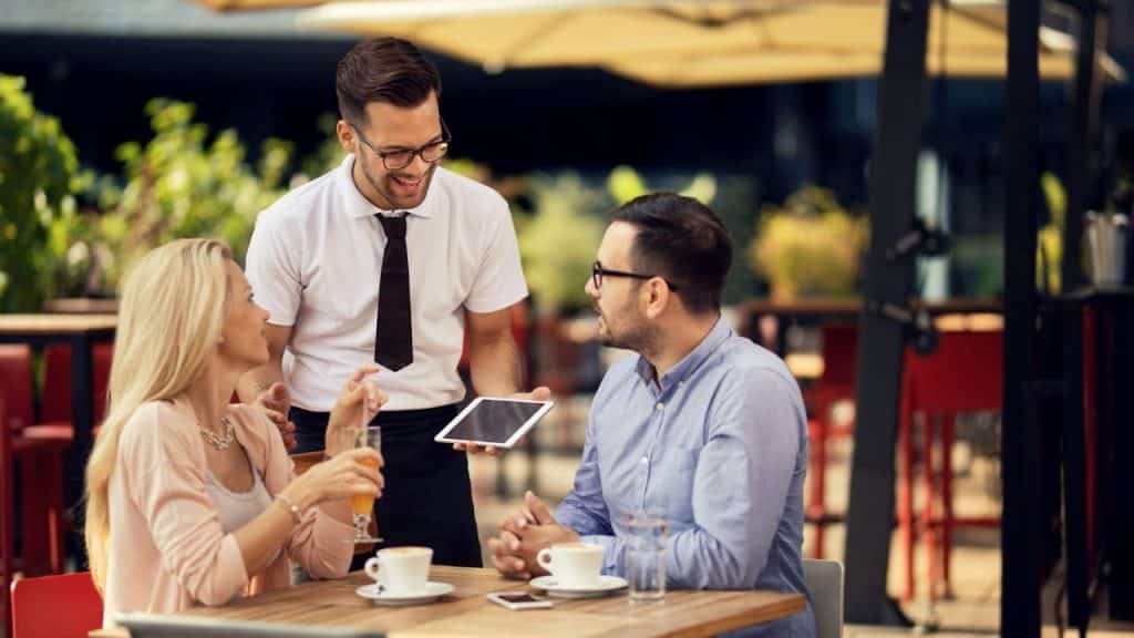A server holding a tablet smiles while taking an order from a casually dressed couple at an outdoor cafe.