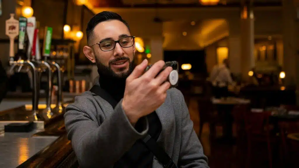 A man with a beard and glasses sits in a dim bar, looking at and holding up his smartphone.