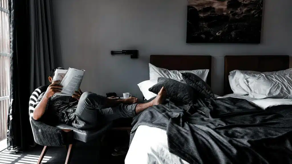A man reading a book while sitting beside an unmade bed.