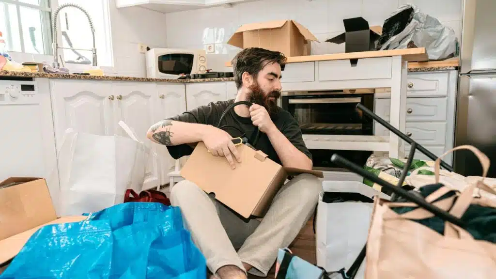 A bearded man sits cross-legged on a kitchen floor surrounded by moving boxes and bags.