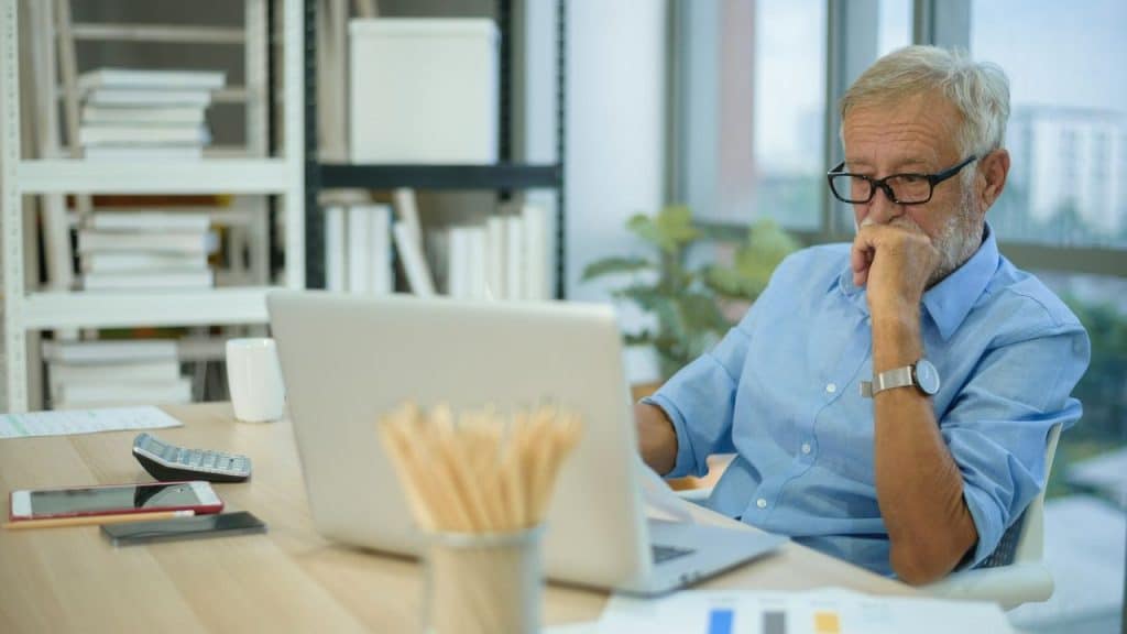 Older man with glasses sitting at a desk and looking intently at a laptop, hand on his chin.