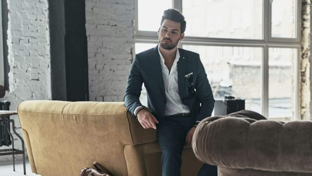 Stylish man in a dark suit leaning against a camel-colored sofa in a bright, modern room.