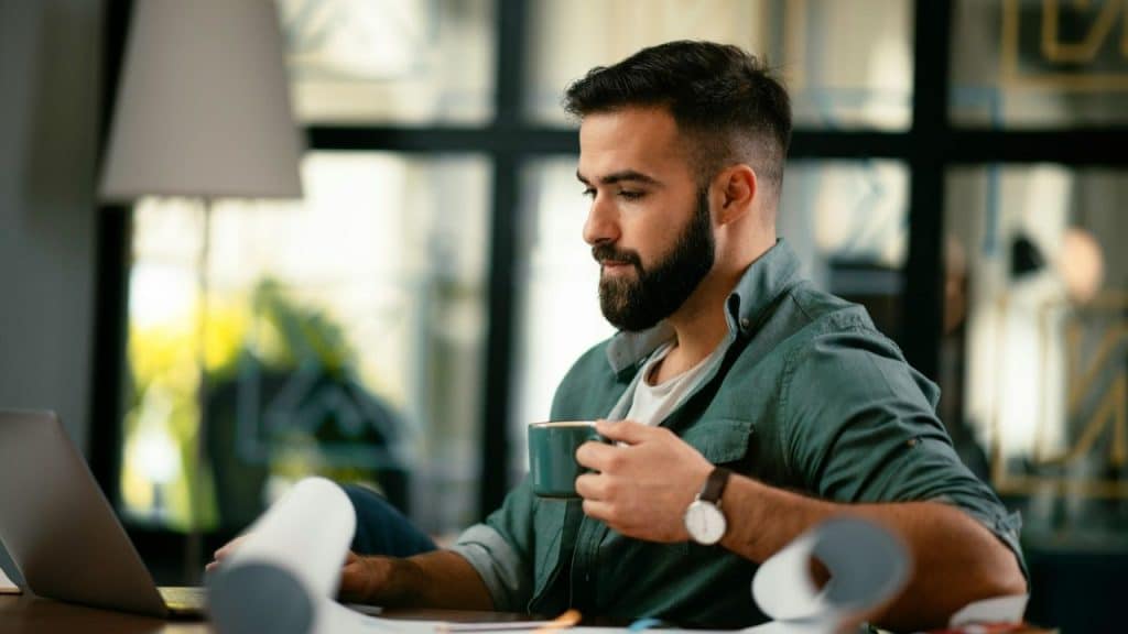 Bearded man working on a laptop while holding a mug in an office setting.