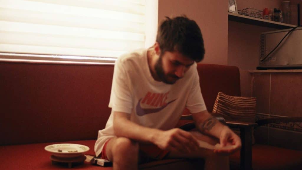 Young man with a beard and tattoo sitting on a red sofa, looking down at his hands.