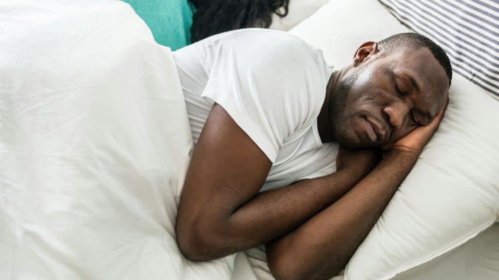 African American man sleeping on his side, under a white blanket and pillow.