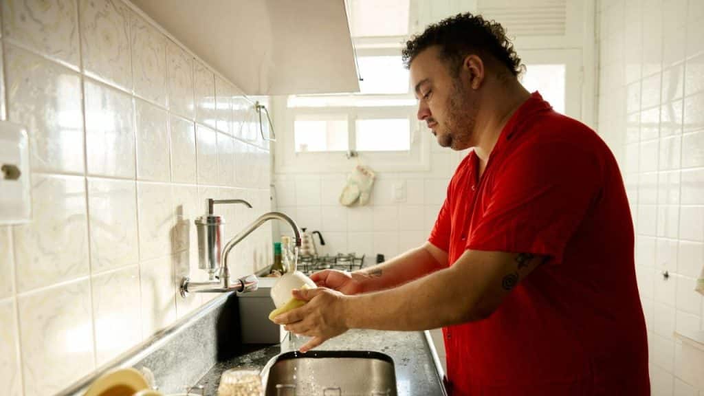 Man in a red shirt with a curly mohawk washing a dish in a kitchen sink.