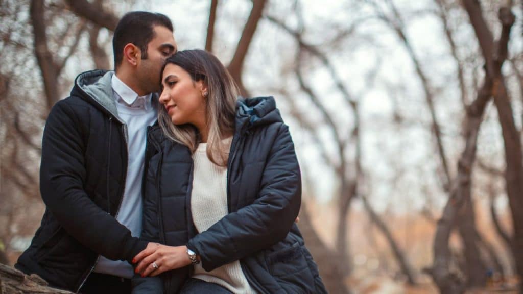 Man wearing a black jacket kissing a woman's forehead in a park with bare trees.