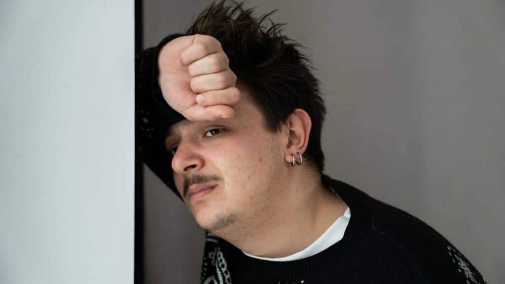 Young man with messy hair and earrings leaning against a wall, resting his forehead on his fist.