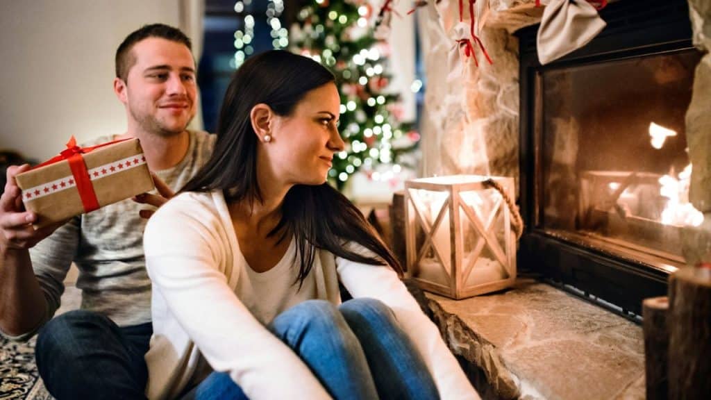Man giving a wrapped gift to a woman by a lit fireplace and Christmas tree.