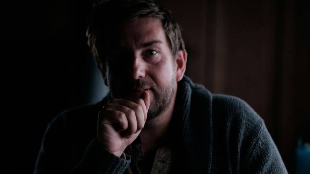 Man in a dark, dramatic light with a beard, hand covering his mouth.