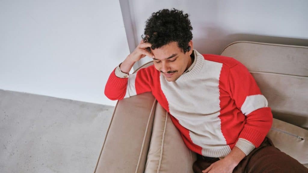 Man with curly hair and a mustache wearing a red and white sweater on a sofa.