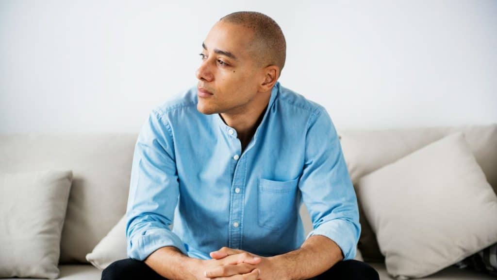 Bald man in a light blue button-down shirt sitting on a beige sofa, looking left.