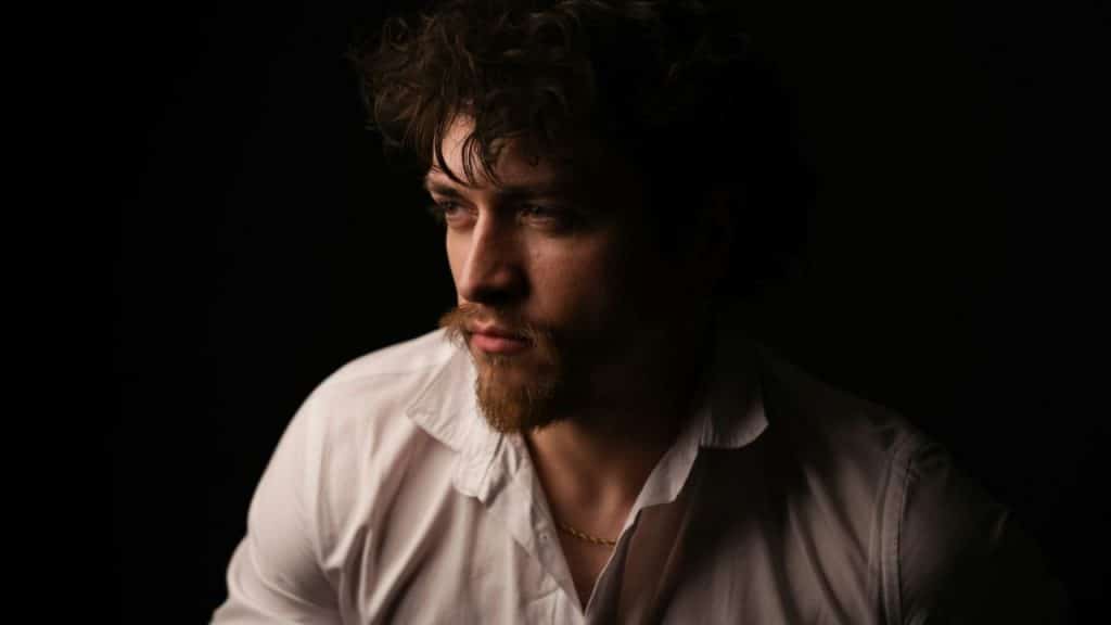 Close-up of a man with curly hair, beard, and mustache, wearing a white shirt.