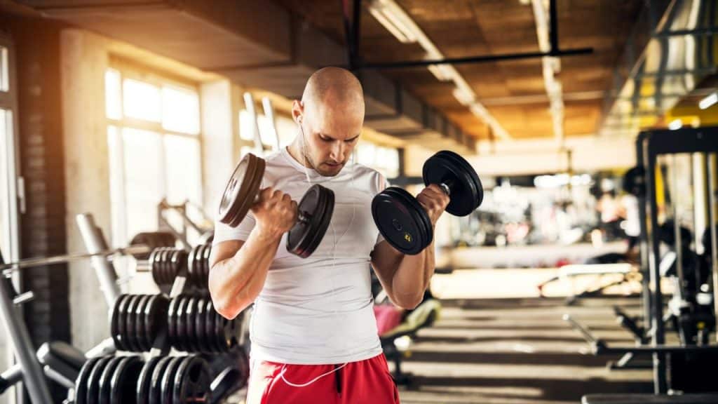 Bald man in a white shirt and red shorts lifting two dumbbells in a sunlit gym.