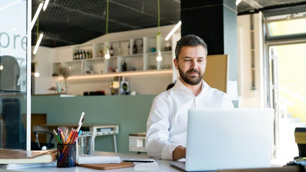 Bearded man in a white shirt working on a laptop at a modern desk in an office.