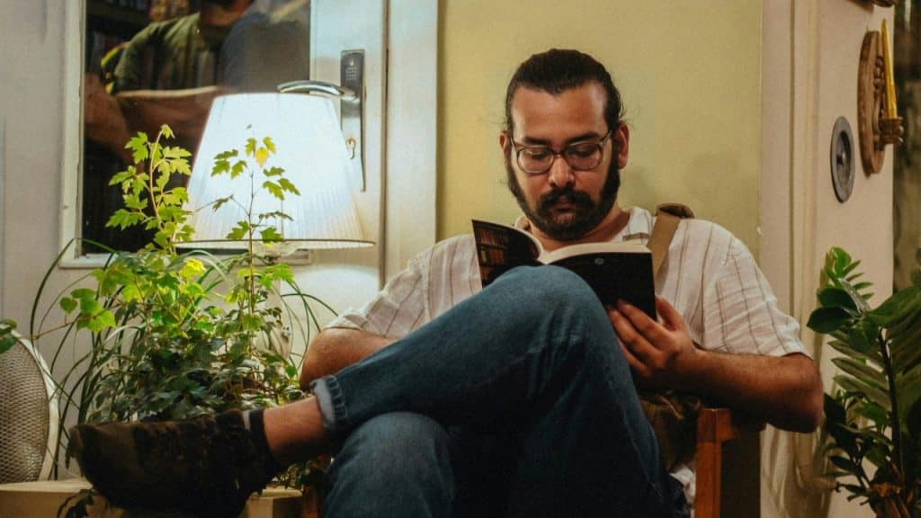 Man with glasses, beard, and ponytail sitting with his legs crossed, reading a book.