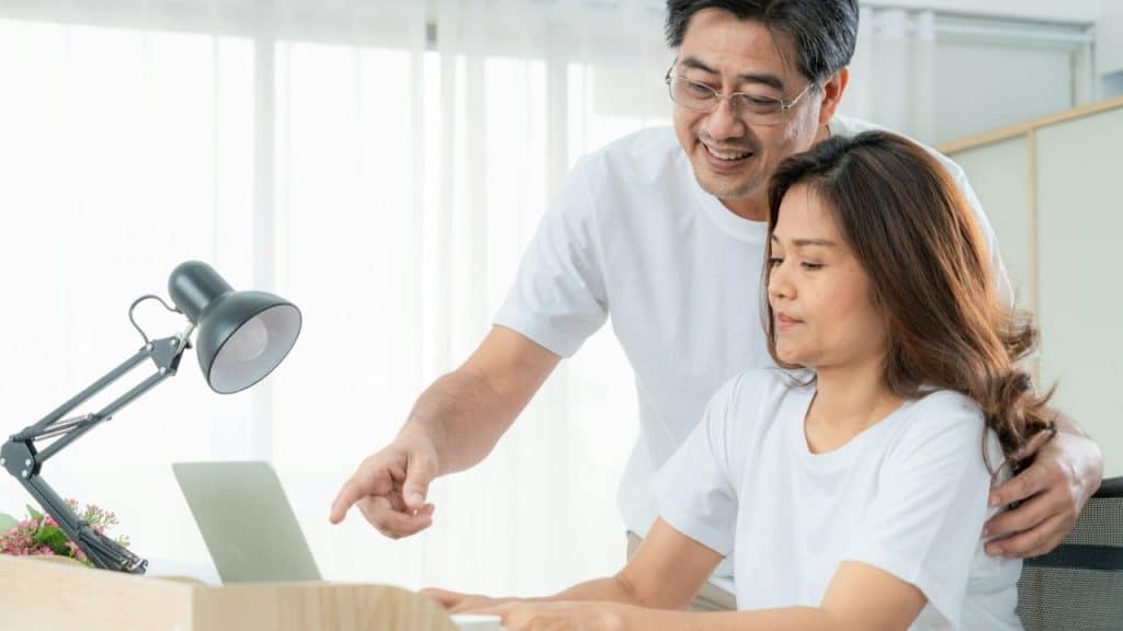 Middle-aged couple in white shirts looking at a laptop, the man pointing at the screen.