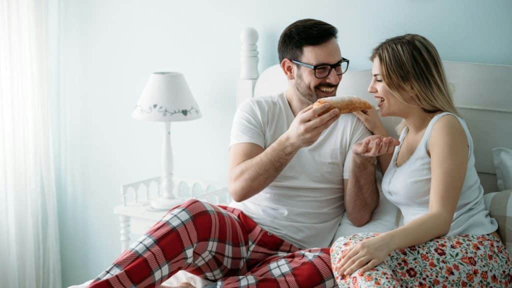 Smiling man in glasses and pajamas feeding a pastry to a woman in bed.