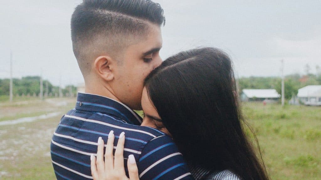 Young man in a striped shirt kissing a woman's forehead as they embrace outdoors.