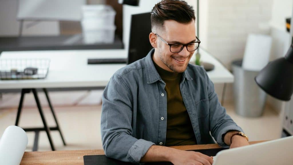 Smiling man with glasses and a beard working on a laptop at a modern desk.