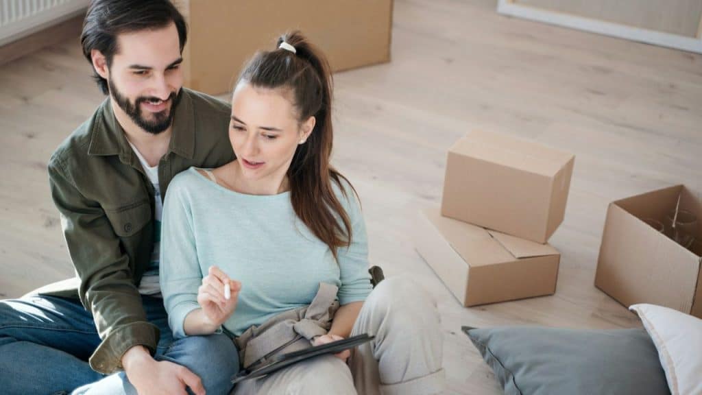 Smiling couple sitting on a wooden floor surrounded by moving boxes, looking at a tablet.