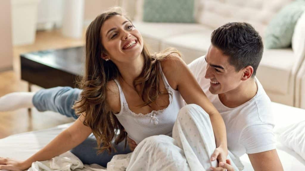Smiling, laughing couple sitting in bed, looking at each other in a bright room.