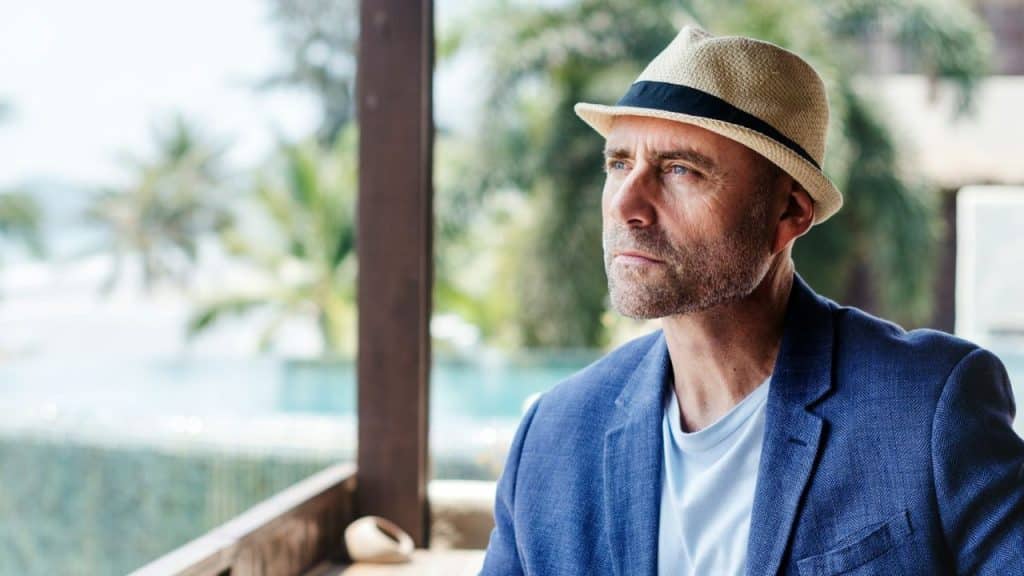 Older man in a fedora and blue jacket looks pensively at a sunny view.