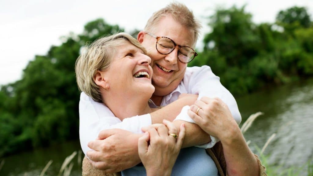Smiling older couple hugging outdoors by a river and green foliage.