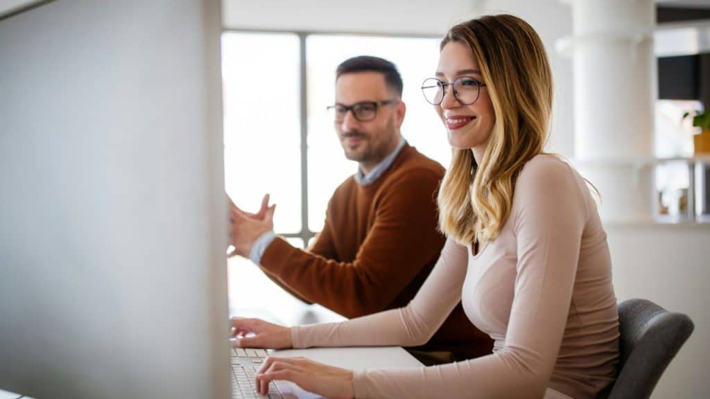 Smiling woman with glasses working at a computer, with a man looking at her.