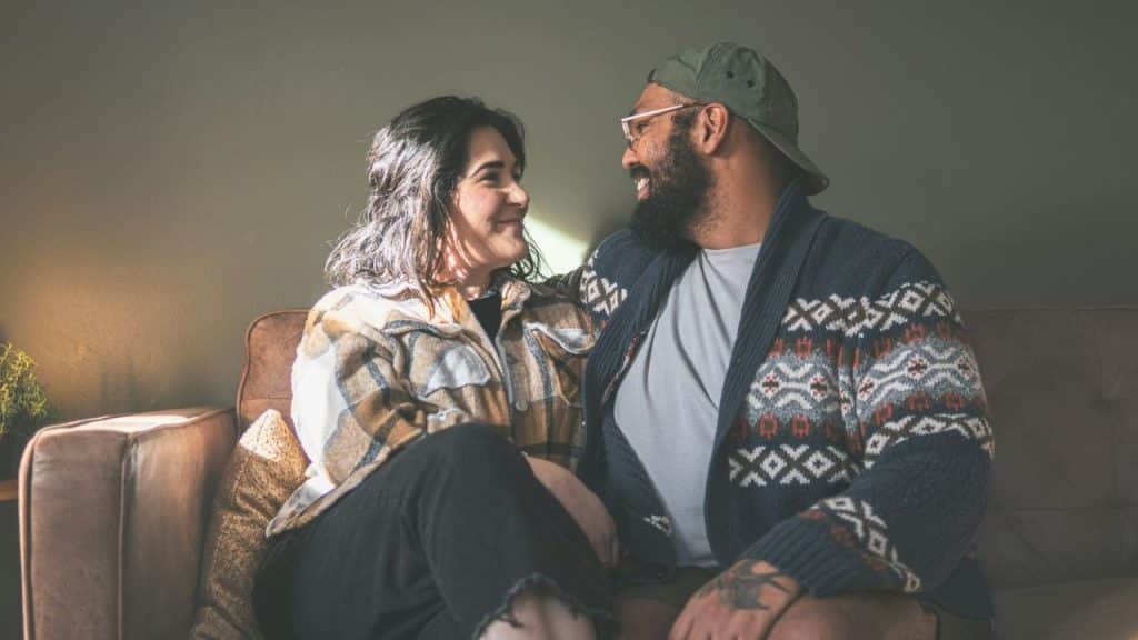 Smiling couple with a beard and glasses sitting on a sofa and looking at each other.
