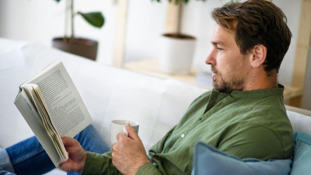 Man in a green shirt sitting on a white couch, reading a book and holding a mug.