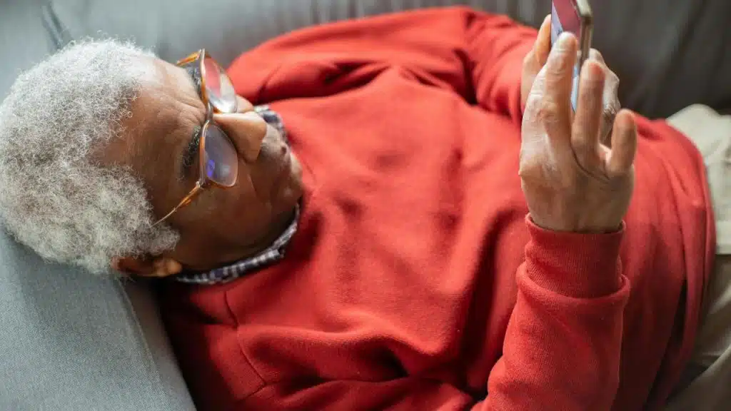 A senior man lying on a couch looking at his phone.