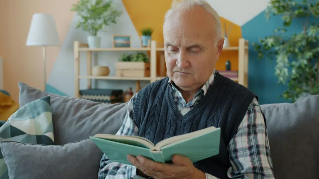 An older man in a knit vest and plaid shirt sits on a sofa reading a book.