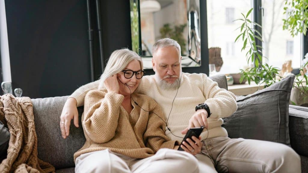 A senior couple sitting on a couch sharing earphones while looking at a phone.