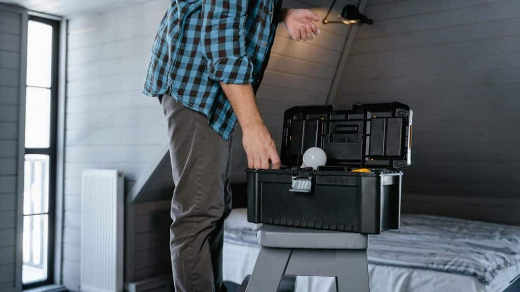 A person standing on a step stool reaching into a toolbox with a light bulb.