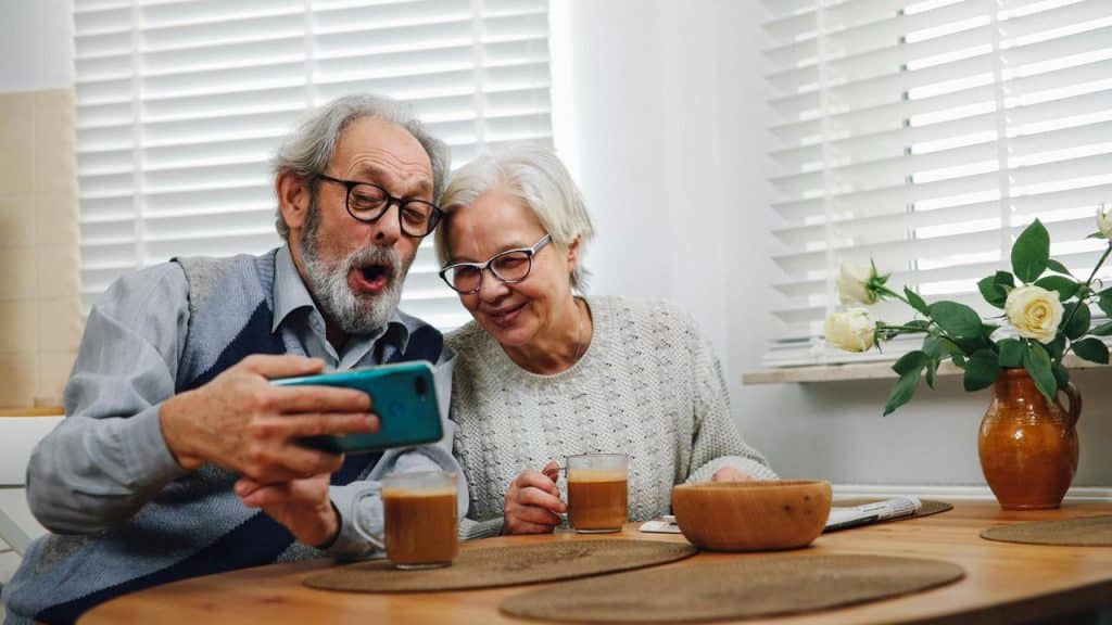 A senior couple sitting at a table smiling while looking at a phone.