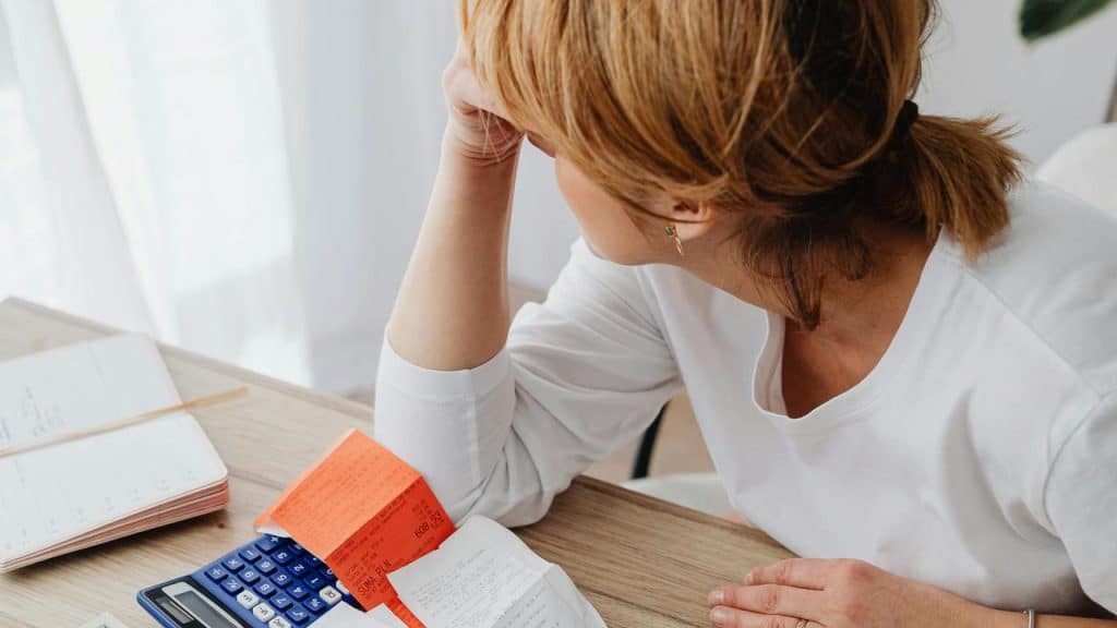 A woman sitting at a table looking stressed while reviewing bills with a calculator.