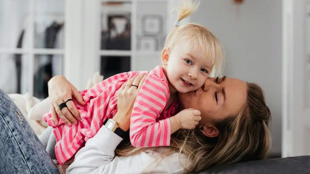 A mother lying on a couch kissing her smiling toddler in a pink striped dress.